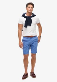 T-shirt bianco, maglione blu navy legato attorno al collo, pantaloni corti azzurri, cintura marrone e scarpe da barca bordeaux. Outfit estivo casual.