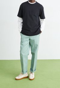 Zwart T-shirt met korte mouwen gedragen over een wit T-shirt met lange mouwen, mintgroene broek en witte sneakers met beige zolen op een groene achtergrond.