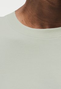 Ανοιχτό πράσινο βαμβακερό t-shirt με στρογγυλή λαιμόκοψη και ομαλή υφή, εμφανίζοντας μέρος του λαιμού και της περιοχής των ώμων.