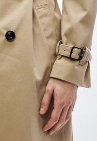 Béžový bavlnený trench coat s čiernym gombíkom, nastaviteľným remienkom na manžete a hladkou textúrou, zdôrazňujúci detaily v oblasti rukáva.