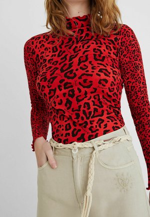 Nainen, joka käyttää punaista ja mustaa leopardikuvioista pitkähihaista paitaa, joka on tungettu beigen värisiin housuihin, joissa on punottu vyö, ja hänellä on käsi taskussa.