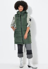 Gilet verde gonfio con colletto alto e zip frontale, dotato di cordini, abbinato a una camicia con motivo a stelle, pantaloni patchwork grigi e neri, e stivali argentati.