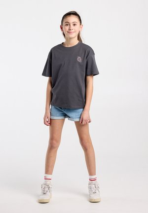 Jeune personne debout portant un t-shirt gris foncé avec un petit motif de fraise, un short en jean, des chaussettes blanches à rayures rouges et des baskets blanches.