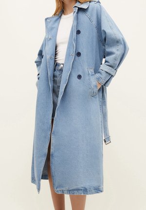 Femme portant un long trench-coat en denim bleu clair sur une chemise blanche et un short en denim, mains dans les poches du manteau, sur fond beige.