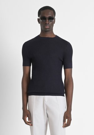 Maglione a maniche corte in maglia a coste di colore blu navy con scollatura rotonda, abbinato a pantaloni chiari, su uno sfondo chiaro.