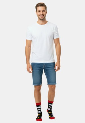 Weißes kurzärmeliges T-Shirt, blaue Jeans-Shorts und schwarze Socken mit bunten Lebensmittelmustern auf rotem Hintergrund. Stehende Haltung.