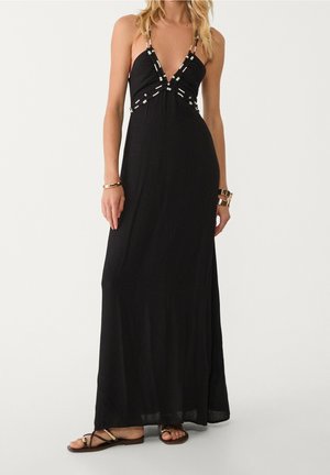 Woman wearing a long black sleeveless dress with beaded detailing on the neckline and waist, paired with flat sandals and gold bracelets.