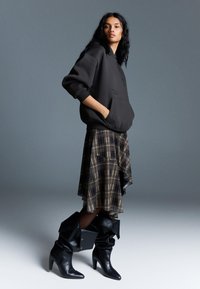 Čierna mikina s kapucňou a predným vreckom, skombinovaná s asymetrickou kockovanou sukňou. K nohám sú čierne kožené čižmy po kolená s prehnutým dizajnom, ktoré dokončujú celý vzhľad.