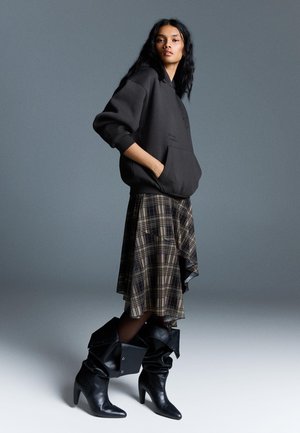 Schwarzer Hoodie mit einer Fronttasche, kombiniert mit einem asymmetrischen Rock aus Karomuster. Knielange schwarze Lederstiefel mit gefaltetem Design runden das Outfit ab.