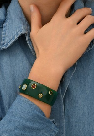Bracelet en résine verte avec des accents dorés, comportant plusieurs incrustations circulaires de couleurs et tailles variées, porté sur un poignet recouvert de denim.
