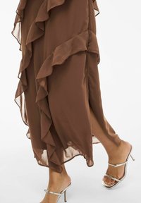 Bruine, vloeiende jurk met gerimpelde randen en een zijsplit, gecombineerd met zilveren sandalen met hak. Lichtgewicht, chiffon-achtige textuur.