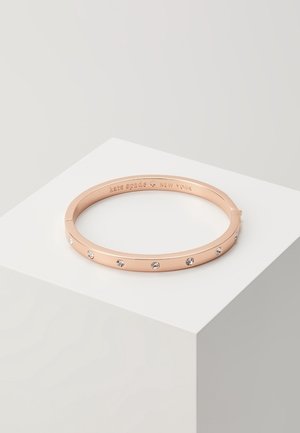 Bracelet jonc en or rose avec des pierres précieuses claires espacées uniformément et "kate spade new york" gravé à l'intérieur, présenté sur un socle blanc.