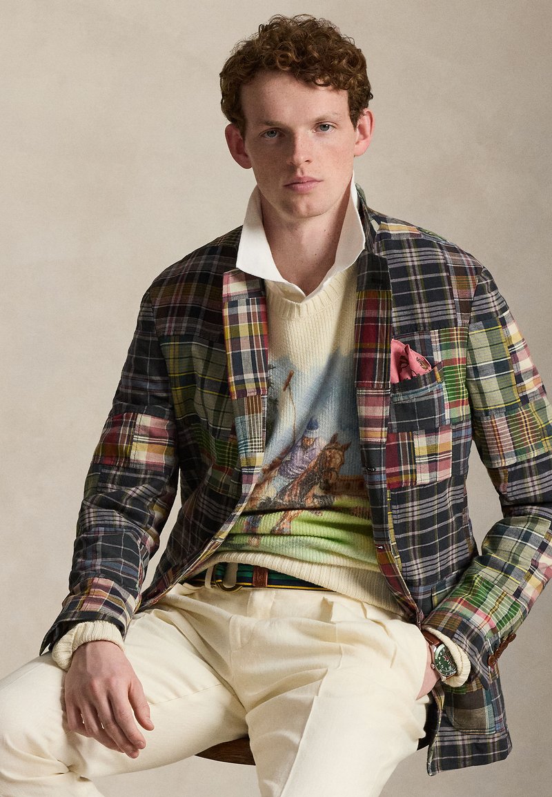 Νέος άνδρας με σγουρά κόκκινα μαλλιά φοράει σακάκι με κολάζ καρό σχέδιο, εικονογραφημένο πουλόβερ με άλογο και ιππέα, λευκό πουκάμισο, κρεμ παντελόνι και πράσινο ρολόι.