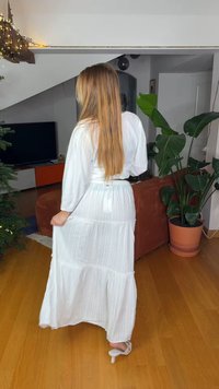 Λευκό φόρεμα μάξι με στρώσεις, μακριά μανίκια και ζώνη με κορδόνι στη μέση. Το ύφασμα είναι ελαφρύ με ρυτιδώδη υφή. Φορεμένο με σανδάλια με τακούνι.