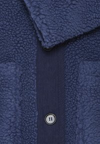 Veste en polaire bleu marine présentant une surface extérieure texturée, des accents côtelés, deux poches et une fermeture à bouton noire.