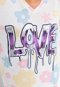 Biała koszulka z dekoltem w serek, z wyraźnym fioletowym napisem "LOVE" z srebrnymi akcentami i tłem w pastelowe wzory kwiatowe.