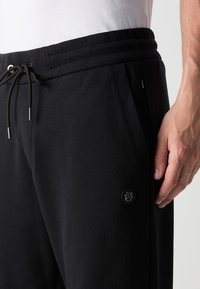 Svarta sweatpants med ribbad midja, dragsko, sidofickor och en logopatch på det nedre benet. Mjuk tygtextur.