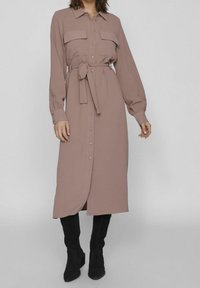 Dlouhá fialová košilová šaty s knoflíkovým zapínáním, límečkem, dvěma náprsními kapsami a páskem v pase. Nosí se s černými kotníkovými botami. Hladká texturovaná tkanina.