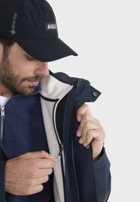 Giacca impermeabile blu marina con fodera in pile bianco, dotata di cerniere argento, un bottone e un cappello nero con il marchio "AIGLE."