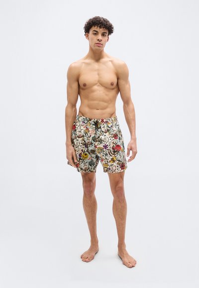 Νέος άνδρας με σγουρά μαλλιά, φορώντας μαγιό με λουλουδάτο μοτίβο, όρθιος ξυπόλητος μπροστά σε μια απλή φωτεινή φόντο.