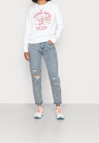Felpa bianca con stampa grafica rossa, jeans distressed azzurri chiari e sneakers bianche con dettagli arancioni e turchesi. Taglio casual.