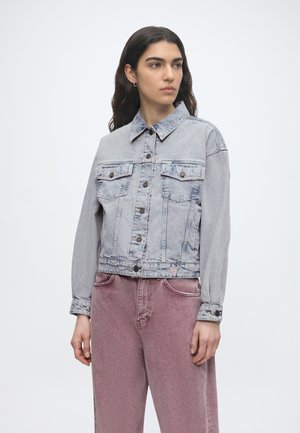 Kvinde med langt mørkt hår iført en lysegrå denimjakke og højtaljede falmede lyserøde jeans, der kigger til siden.