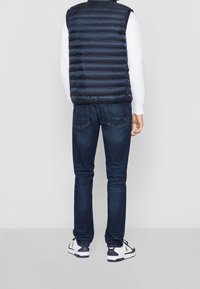 Granatowa puchowa kamizelka z poziomymi paskami, noszona na białej koszuli z długim rękawem. Do tego ciemnoniebieskie dżinsy i białe buty sportowe.