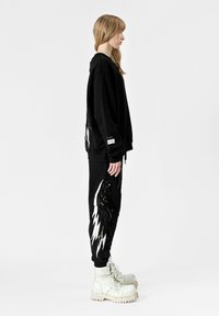 Sudadera negra de gran tamaño con diseños de alas en blanco y dorado, combinada con pantalones de chándal negros con acentos similares, llevada con botas de color claro.
