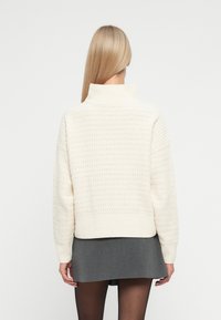 Pull à col roulé crème avec un motif de point texturé. Design court avec un ourlet côtelé. Associé à une mini-jupe grise et des collants noirs.