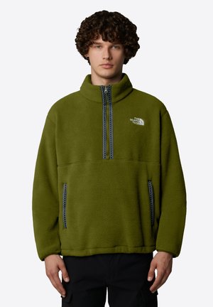 Junger Mann trägt einen olivgrünen Fleecepullover mit halbem Reißverschluss und Taschen vorne, mit dem The North Face-Logo auf der Brust.