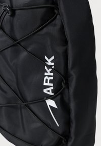 Zaino nero con un tessuto testurizzato e un design con corda elastica sulla parte anteriore, con un logo bianco "ARKK" in evidenza.
