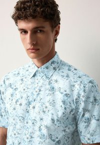 Camicia azzurra a maniche corte con un motivo floreale in diverse tonalità di blu. Presenta un colletto classico e chiusura con bottoni. Tessuto liscio.