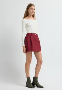 Witte geribbelde langarmtop gecombineerd met een gestructureerde burgundy minirok met plooien en een knoop aan de voorkant. Gekenmerkt door olijfgroene enkellaarzen.