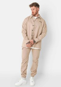 Chaqueta de camisa beige de gran tamaño con diseño de botones, logo en el pecho y puños relajados. Combinada con pantalones beige ajustados y zapatillas blancas.