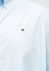 Ανοιχτό μπλε πουκάμισο με λεπτές ρίγες και μικρό ορθογώνιο λογότυπο σε κόκκινο, λευκό και ναυτικό μπλε κεντημένο στο στήθος.