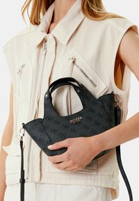 Mujer con chaleco sin mangas beige sosteniendo un pequeño bolso negro de Guess con logo y correa desprendible frente a un fondo blanco.