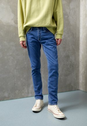 Person, die ein hellgrünes Langarmshirt, blaue Slim-Fit-Jeans und weiße Canvas-Sneaker trägt, steht auf grauem Boden vor einer Betonwand.