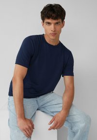 Granatowa koszulka polo z krótkim rękawem, wykonana z miękkiego materiału, z dekoltem w kształcie łódki. Połączona z jasnoniebieskimi dżinsami, model siedzi na białym bloku.