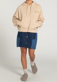 Beige hættetrøje med frontlomme, kombineret med en denimnederdel med to blå nuancer og knapdetaljer, samt grå slip-ons med sokker.