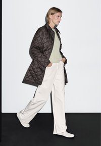 Hnědý prošívaný kabát přes světle zelenou košili, doplněný bílými širokými kalhotami a světlou obuví. Hladké textury a kontrastní barvy.