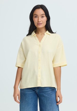 Joven mujer con cabello largo y oscuro, vistiendo una camisa amarilla pálida de manga corta abotonada y jeans azules, de pie frente a un fondo liso.