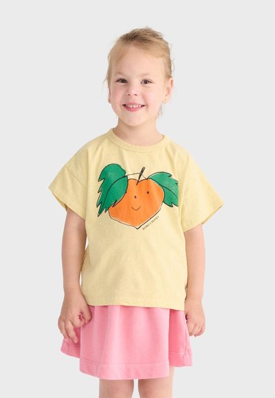 Lächendes junges Mädchen, das ein gelbes T-Shirt mit einer Illustration einer Orange und grünen Blättern sowie einen rosa Rock trägt, vor einem schlichten Hintergrund.