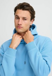 Felpa con cappuccio azzurro chiaro con polsini a coste e cordino. Tessuto morbido con vestibilità comoda. Le mani stanno sistemando il colletto.