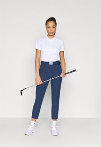 Σετ γκολφ με λευκό πόλο πουκάμισο, σκούρο μπλε παντελόνι cropped και λευκά αθλητικά παπούτσια, κρατώντας ένα μπαστούνι γκολφ με μαύρο και γκρι κοντάκι.