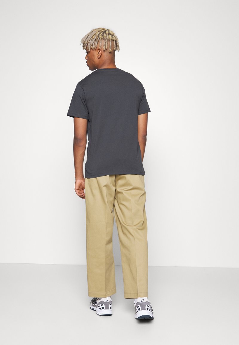 T-shirt in cotone nero, vestibilità rilassata, abbinato a pantaloni beige a gamba larga. Sneakers con dettagli bianchi completano l'outfit. Sfondo bianco.