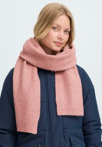 Blondide juustega naine, kes kannab paksu roosa kootud salli ja tumesinist tikkimist jakki, vaatab ettepoole kerge naeratusega.