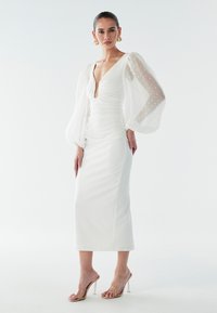 Vit klänning med djup urringning, rynkad design och genomskinliga puffärmar med prickmönster. Matchas med klara högklackade sandaler.