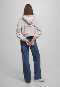 Light pink lightweight jacket with a hood, featuring a relaxed fit and elastic cuffs, paired with loose blue denim jeans and white shoes.