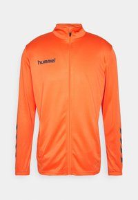 Oranje sportieve ritsjack met hoge kraag, zwart logo op de borst en zwarte diagonale strepen op de mouwen. Gladde, lichte stof.
