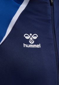 Námornícky športový sako s modro-bielymi akcentmi, so výrazným bielym logom "hummel" vpredu, vyrobené z hladkého, elastického materiálu.
